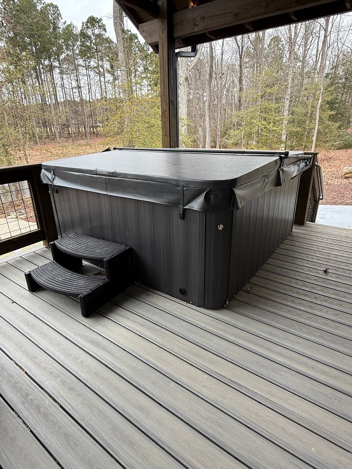 Hot tub covered with a black lid on a wooden deck surrounded by trees.