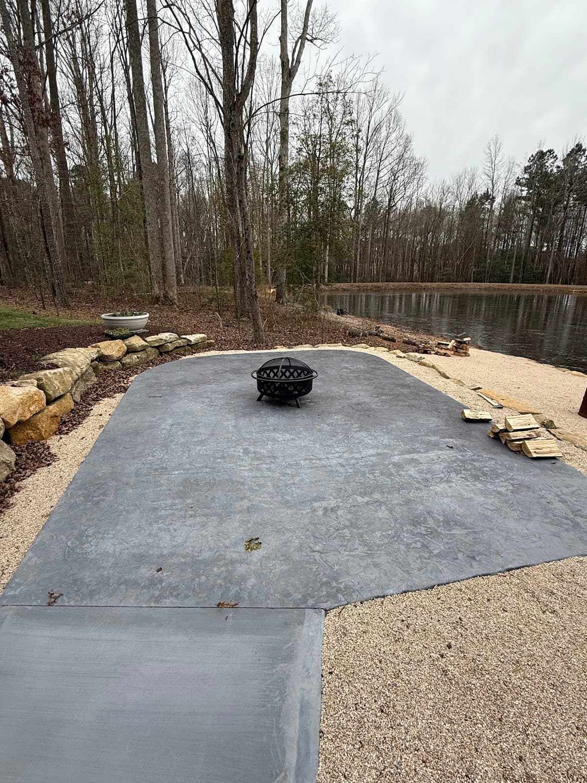 A gray patio area with a fire pit, surrounded by trees and a pond in the background.