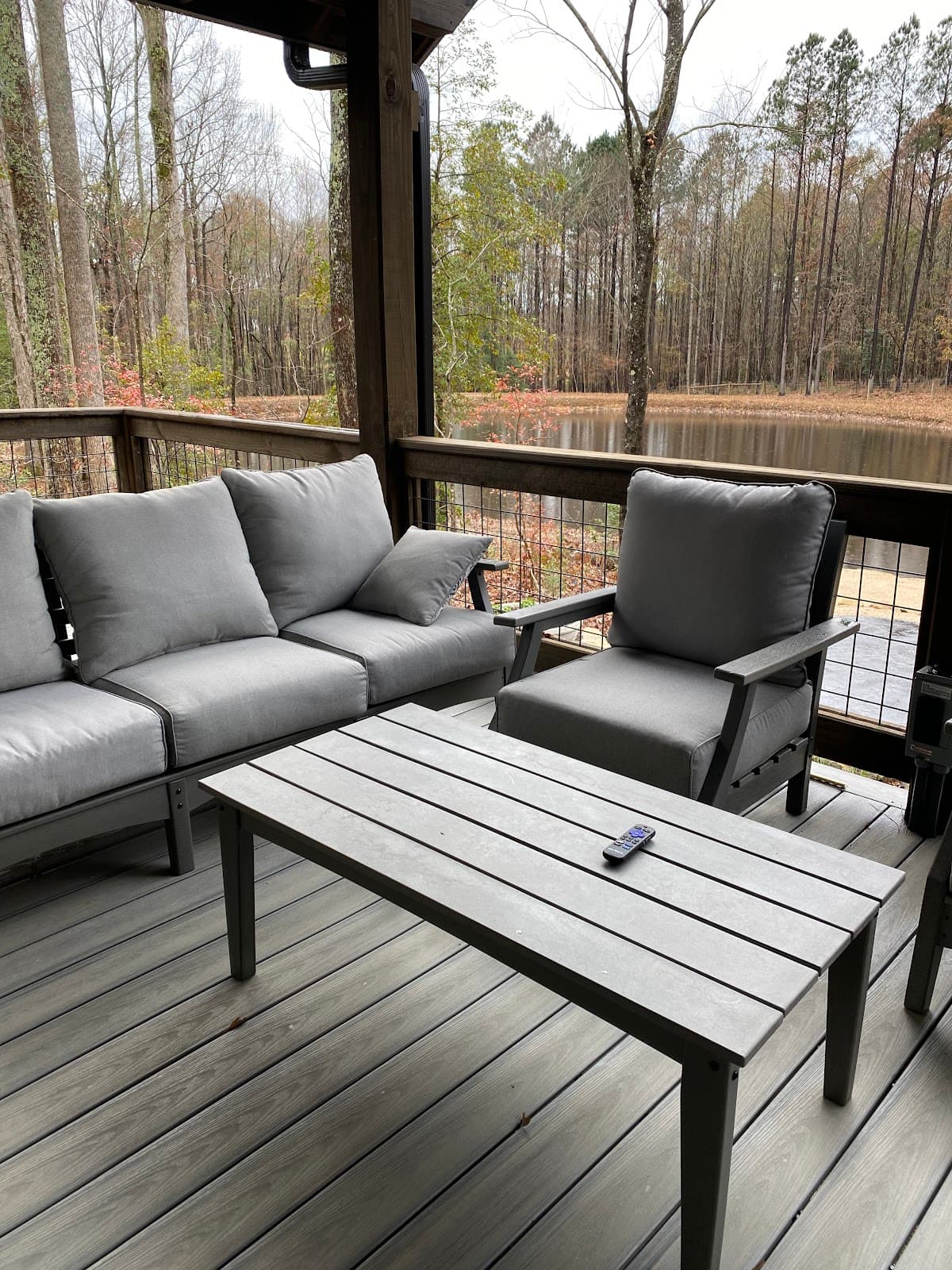 A cozy outdoor seating area with gray cushions overlooks a serene pond surrounded by trees.